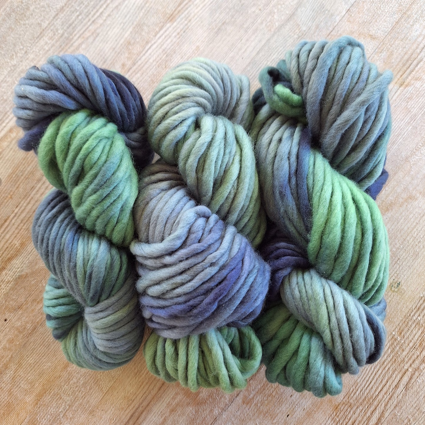 Three skeins of green and blue pencil roving yarn on a wooden surface