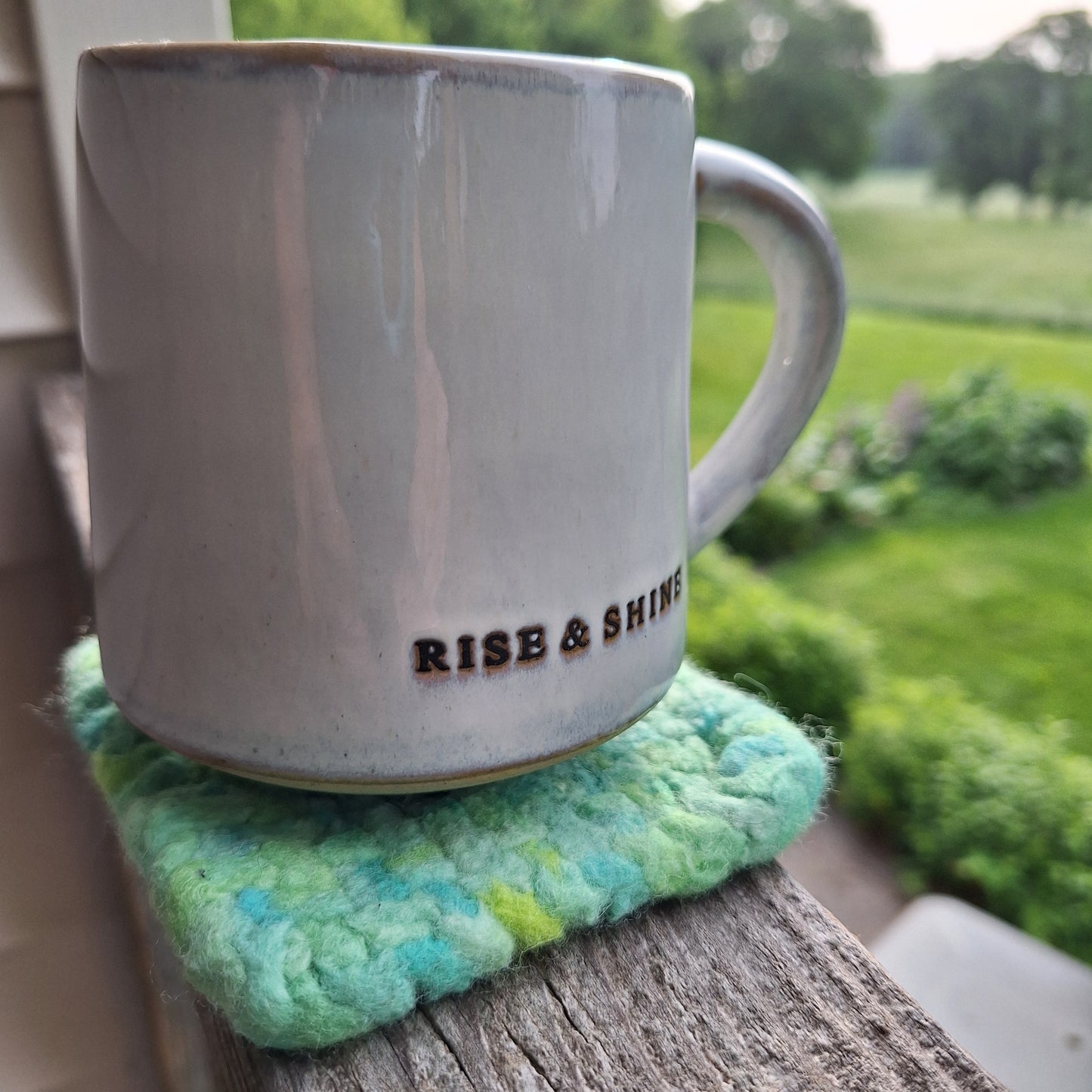 White mug with 'Rise & Shine' text on a green felted wool coaster.