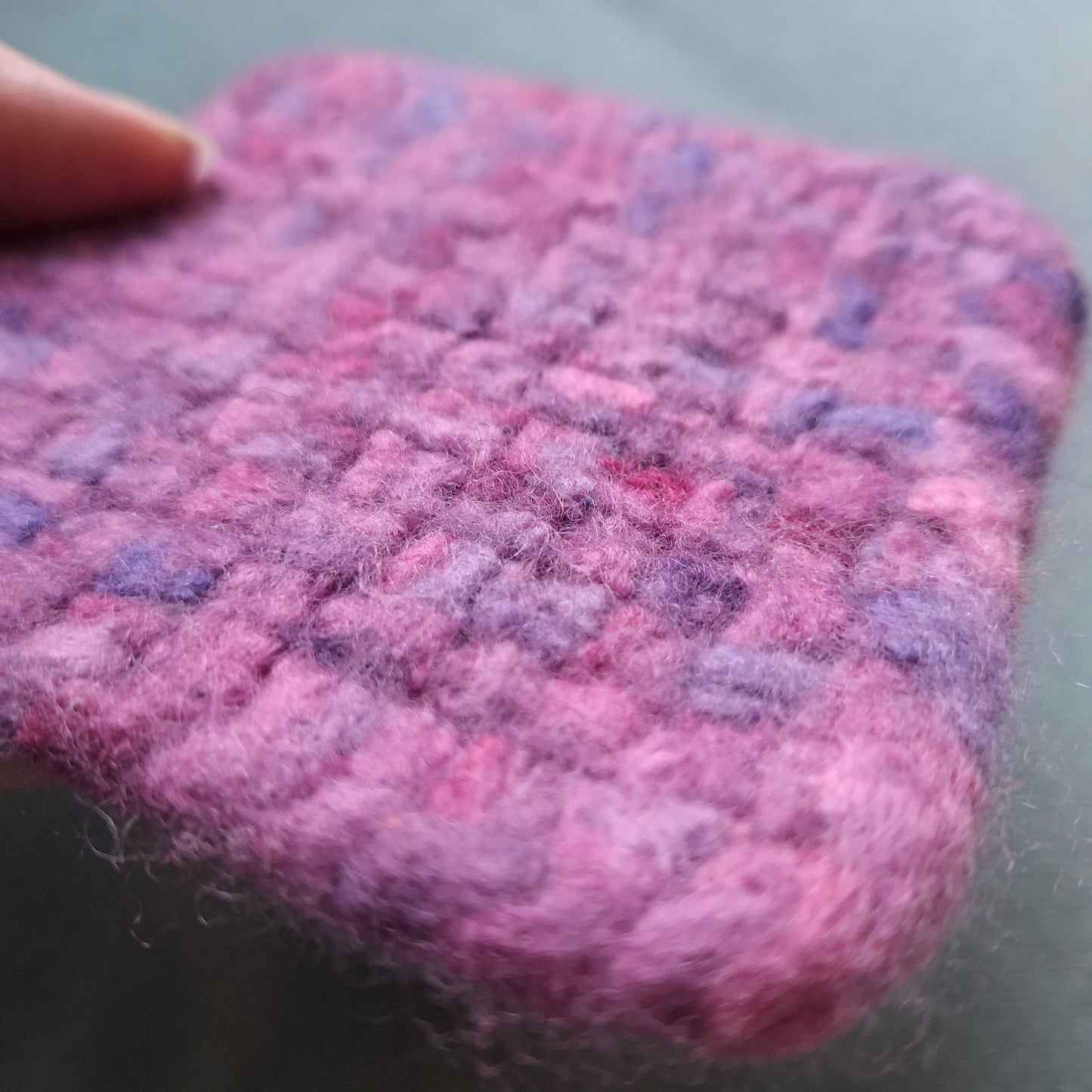 Close-up of a textured pink and purple felted coaster held by a hand against a neutral background