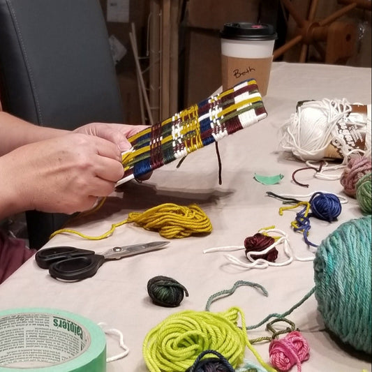 Student working on a weaving at a table in a workshop setting.