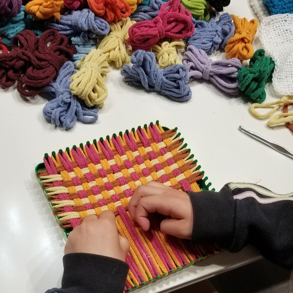 Student working on a potholder weaving project with colorful loops on a table.