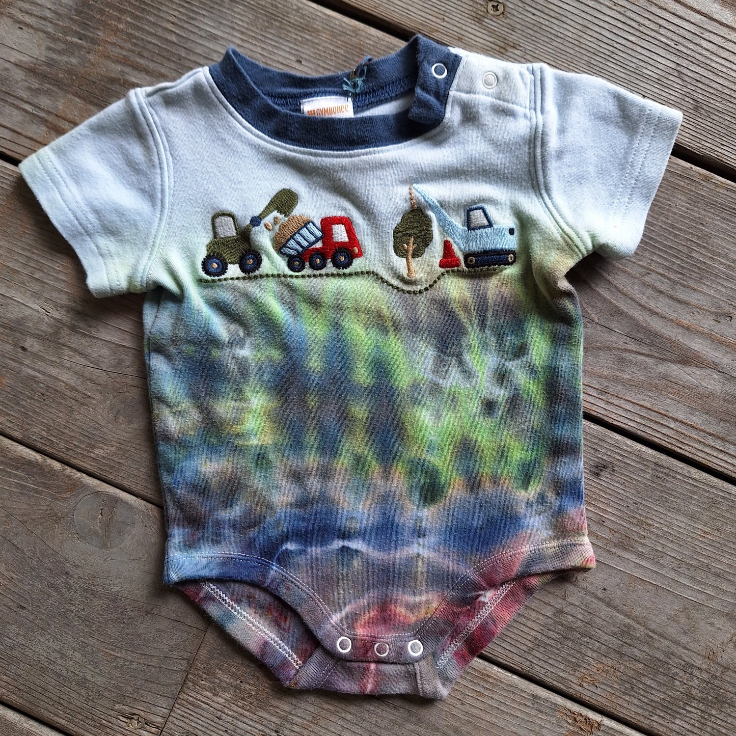 Baby onesie with tie-dye design and toy truck graphics on a wooden surface