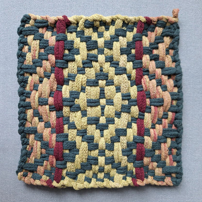 A square woven potholder with a geometric pattern in leaf, pine, variegated ochre, and burgundy, laid flat on a neutral gray background.