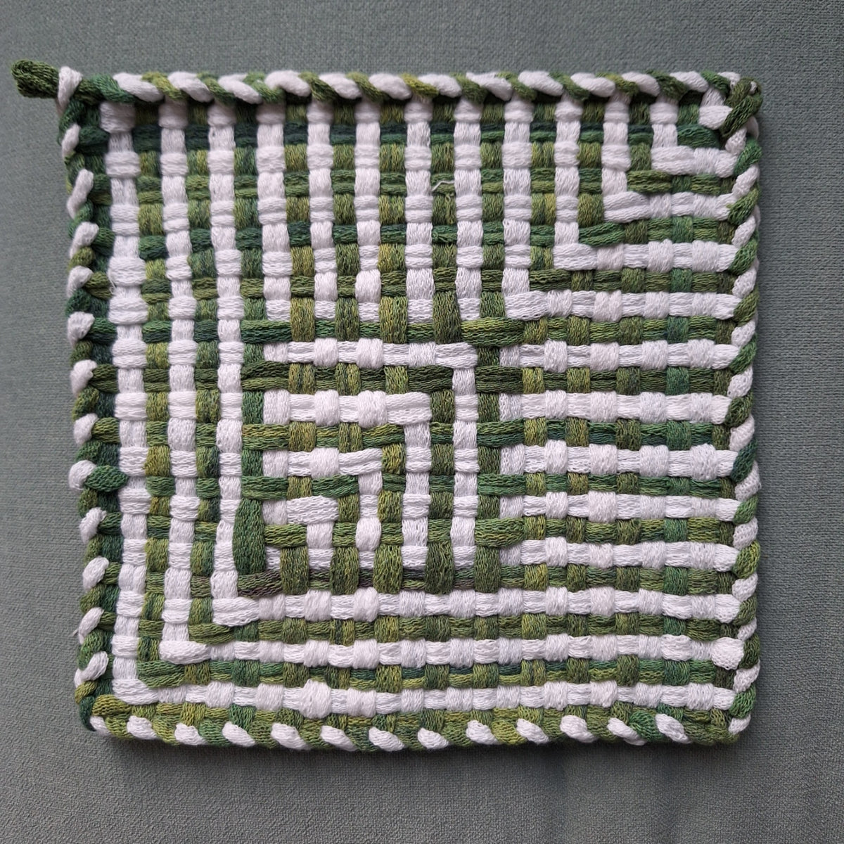 A square woven textile pot holder, featuring a geometric shadow weave pattern in alternating shades of green and white loops, finished with a braided border, shown flat against a neutral gray background.