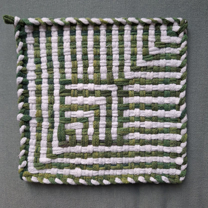 A square woven textile pot holder, featuring a geometric shadow weave pattern in alternating shades of green and white loops, finished with a braided border, shown flat against a neutral gray background.