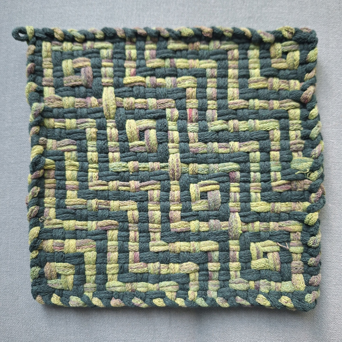 A square woven fabric potholder or mat featuring a geometric maze-like pattern in dark green and light yellow-green loops, with an interlaced border, shown on a neutral gray background.