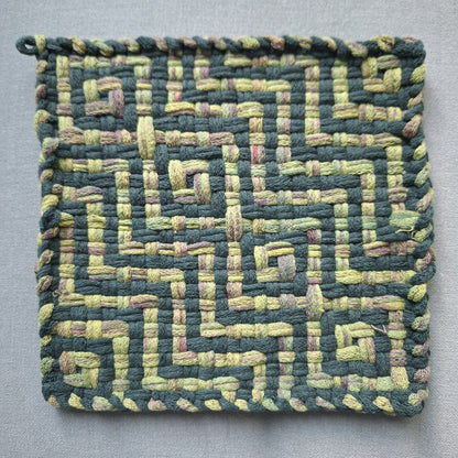 A square woven fabric potholder or mat featuring a geometric maze-like pattern in dark green and light yellow-green loops, with an interlaced border, shown on a neutral gray background.