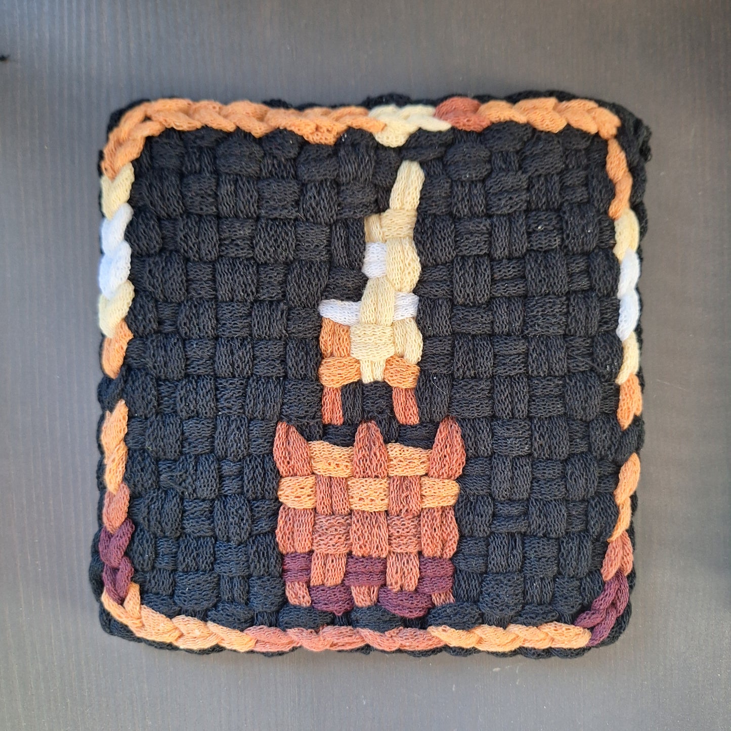 Woven potholder with a candle image and multicolored border on a gray background