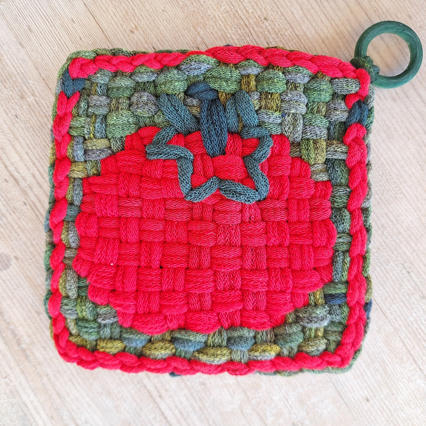 Multicolored woven potholder with a tomato graphic and green ring on a light wooden surface