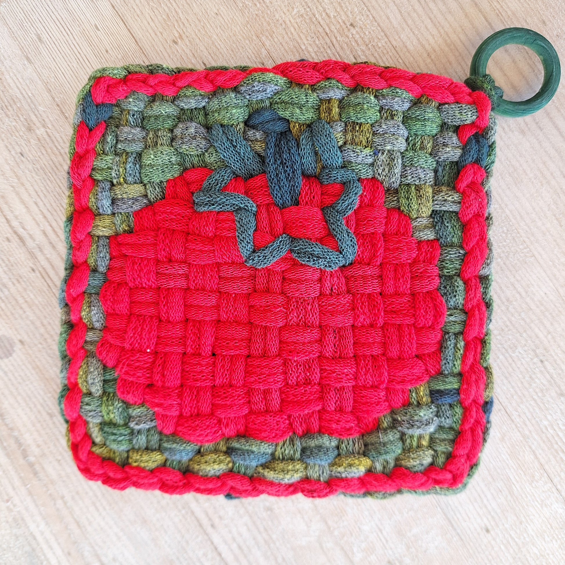 Multicolored woven potholder with a tomato graphic and green ring on a light wooden surface