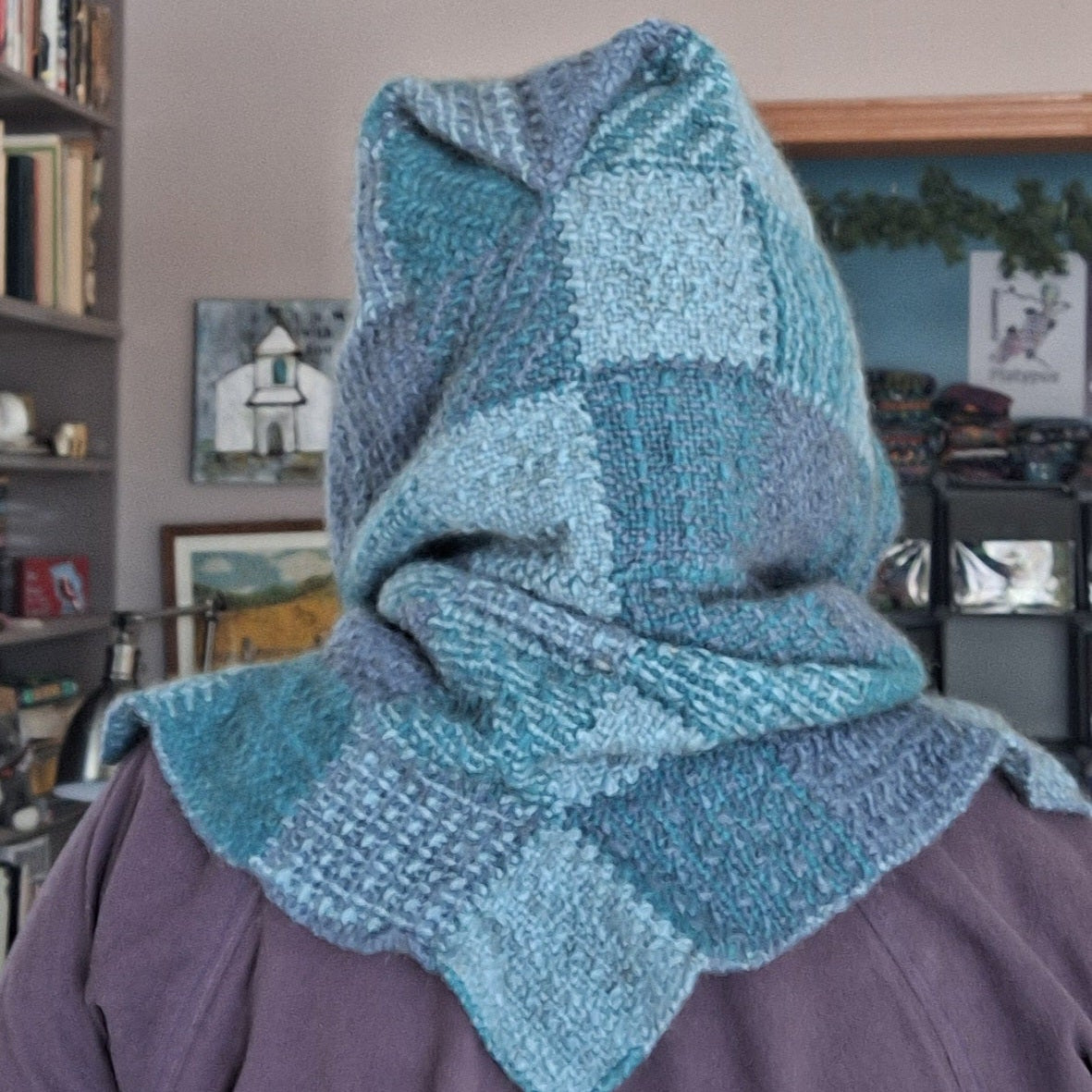 Person wearing a blue patchwork woven hood in a room with books and decor.