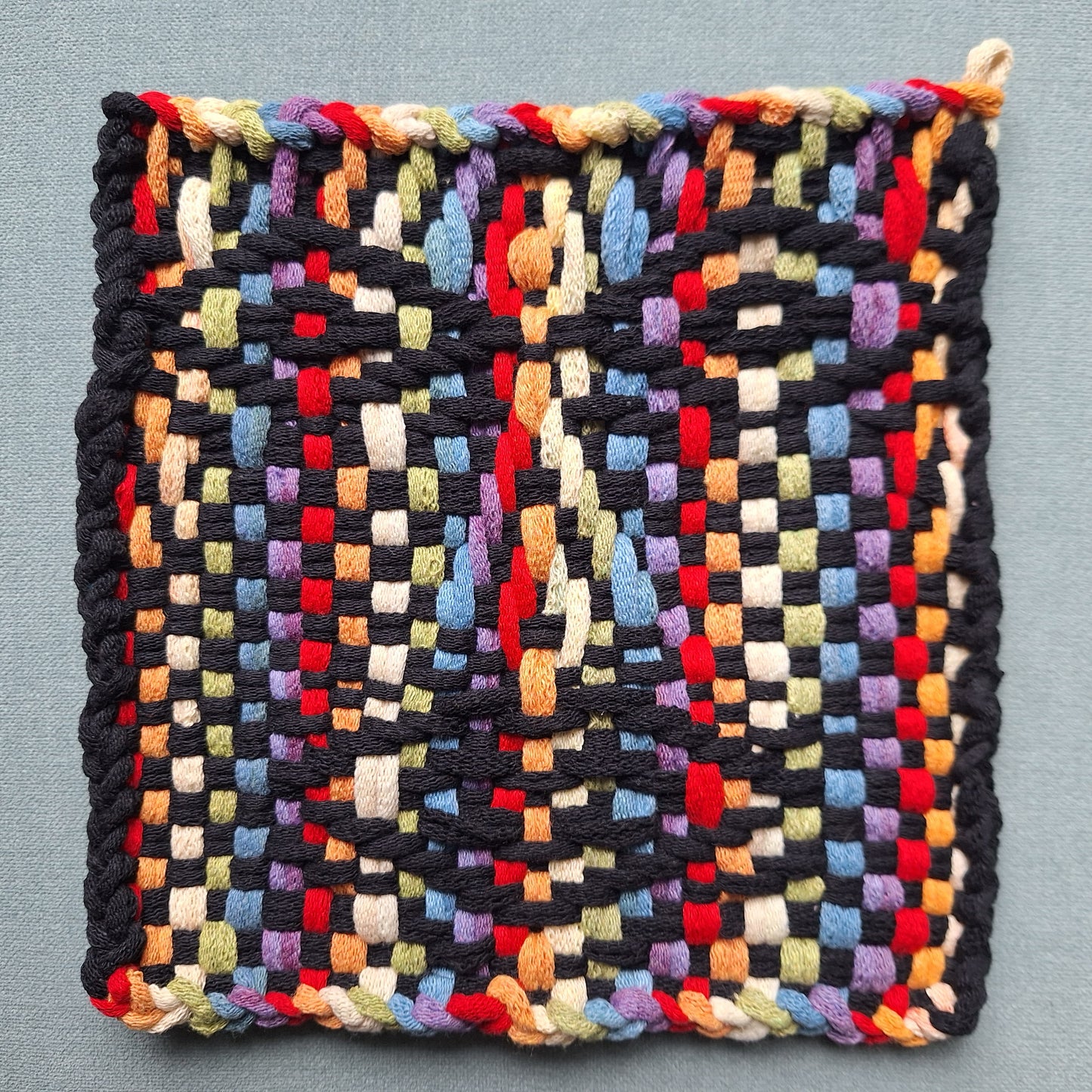Rainbow colored woven square with a face graphic on a gray background