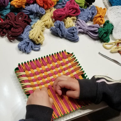 Student working on a potholder weaving project with colorful loops on a table.