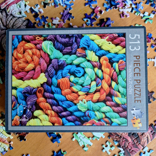 Jigsaw puzzle packaging with colorful yarn design and '513 Piece Puzzle' text on a wooden surface.