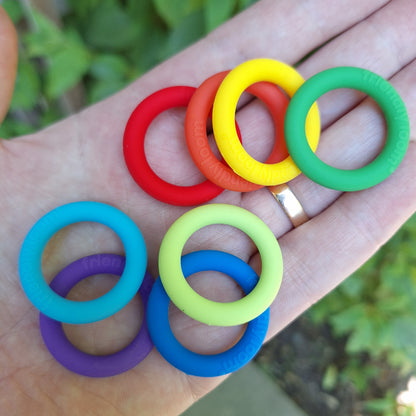 Eight colorful silicone potholder rings in green, yellow, red, lime, green, blue, turquoise, and purple, held in a person's hand with a blurred outdoor garden background.
