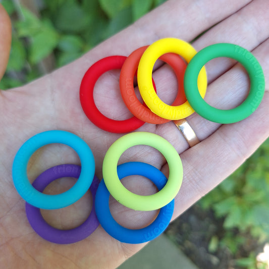 Eight colorful silicone potholder rings in green, yellow, red, lime, green, blue, turquoise, and purple, held in a person's hand with a blurred outdoor garden background.