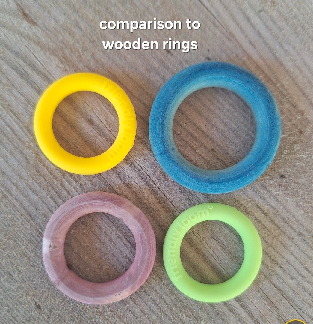 Two circular silicone rings in yellow and lime green and two circular wooden rings in blue and pink, arranged on a light wood grain surface. 