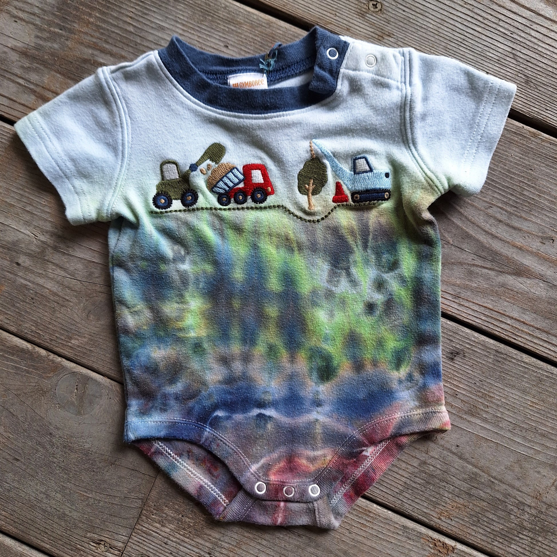 Baby onesie with tie-dye design and toy truck graphics on a wooden surface
