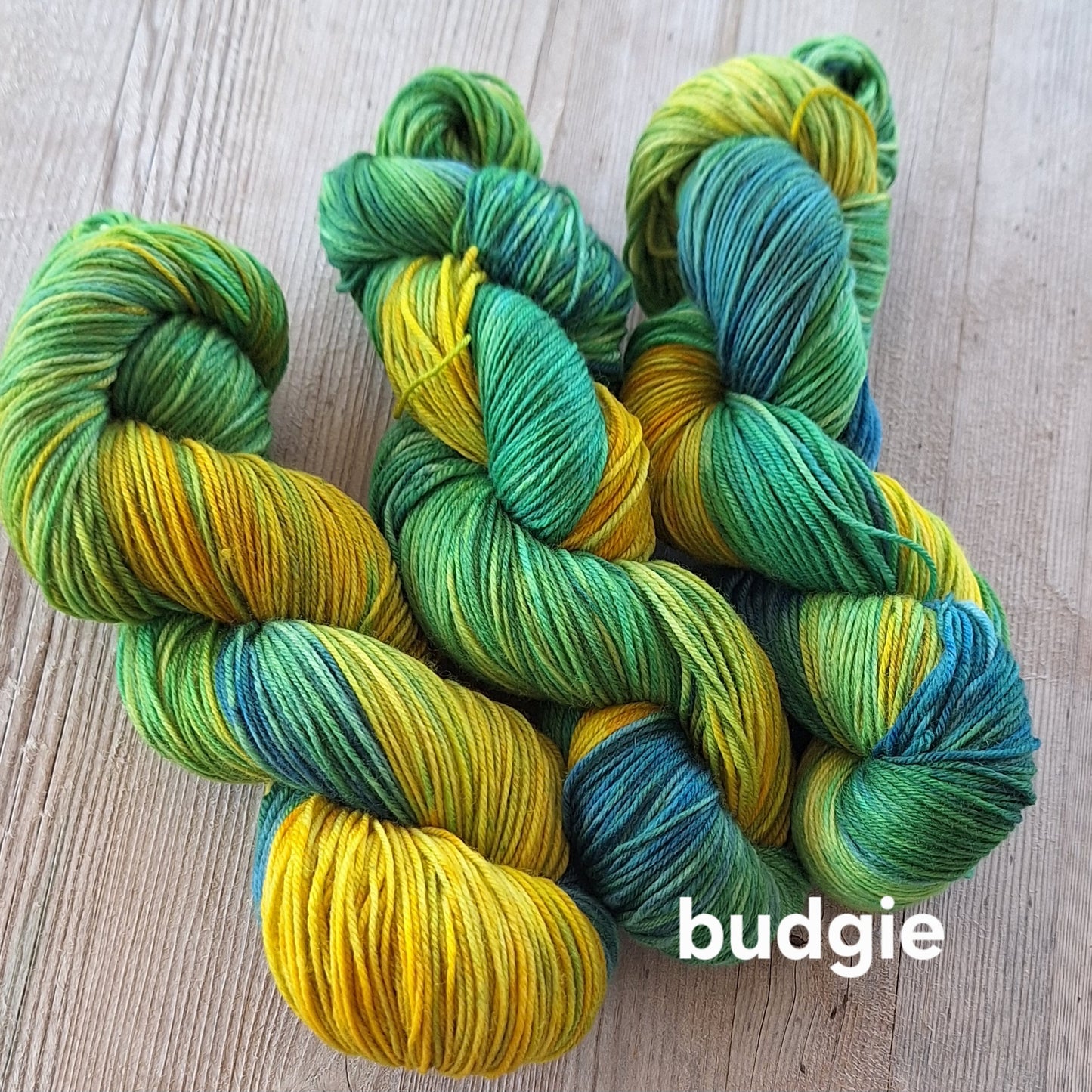 Green and yellow skeins of yarn on a wooden surface with 'budgie' label.