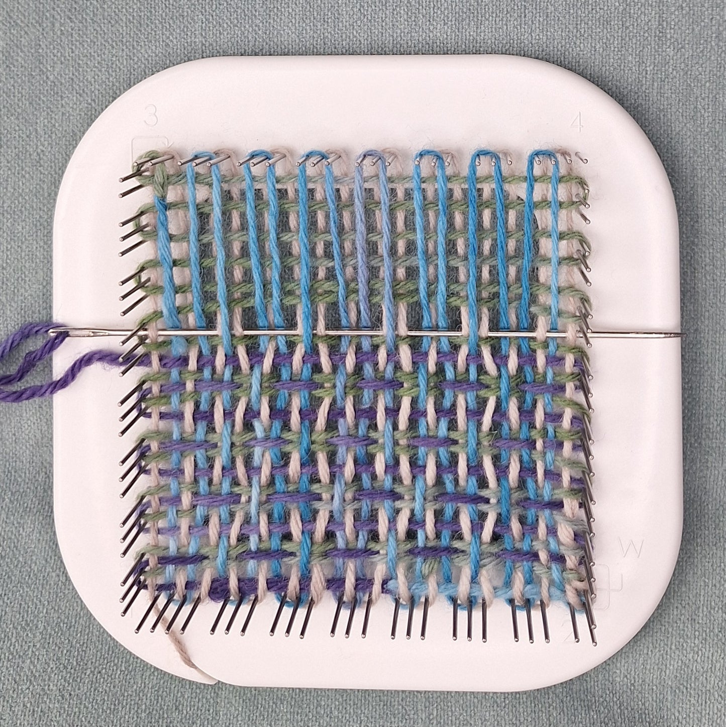 Multicolor weaving project with blue and green yarn on a white Zoom Loom against a gray background