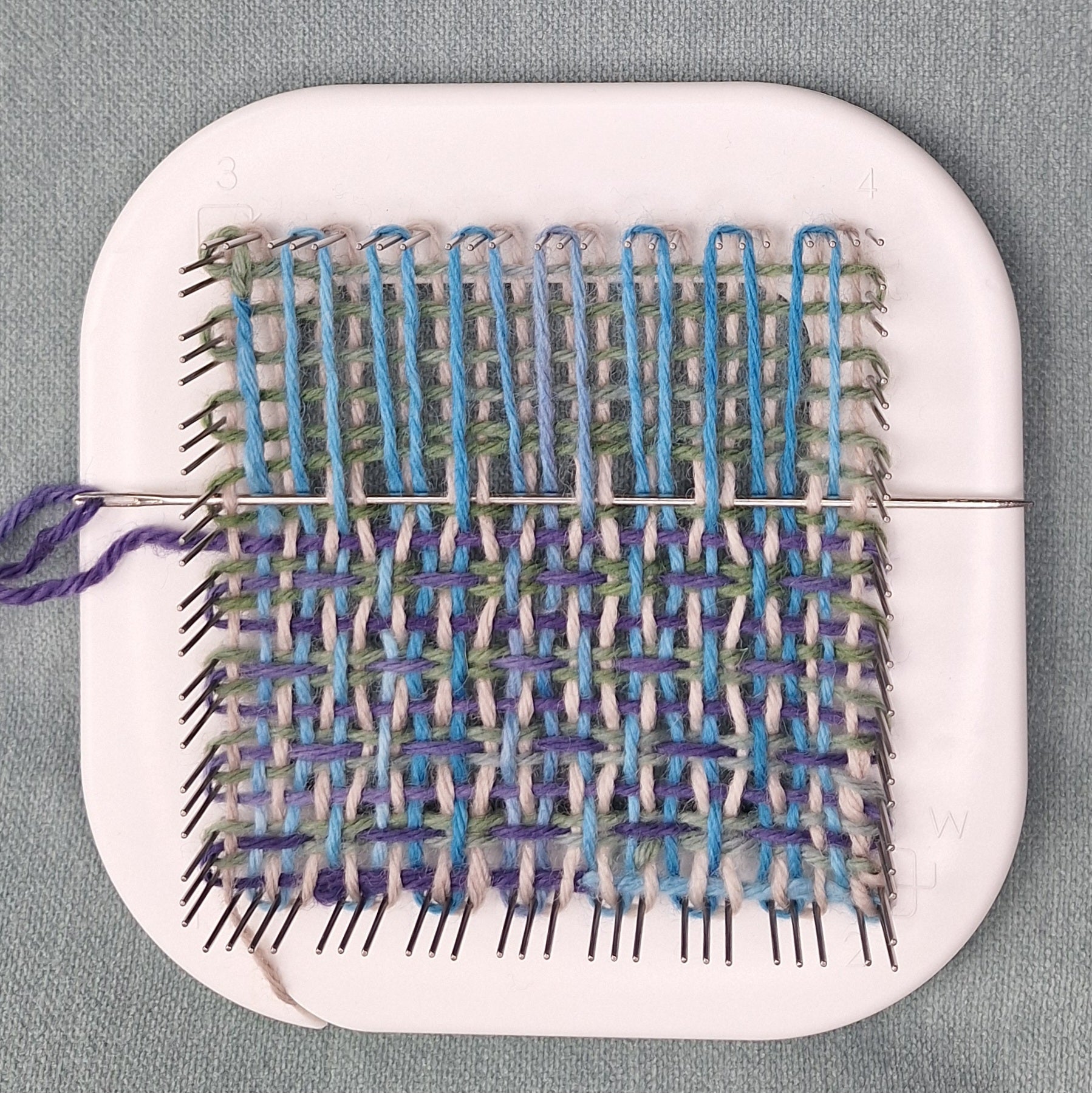 Multicolor weaving project with blue and green yarn on a white Zoom Loom against a gray background